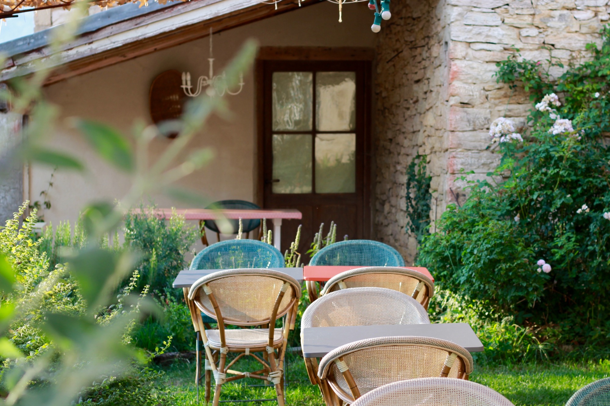 Au fond, l'angle d'une maison en pierre. Au premier plan, quelques tables et chaises qui constituent la terrasse du restaurant, dans une ambiance arborée avec de la pelouse au sol.