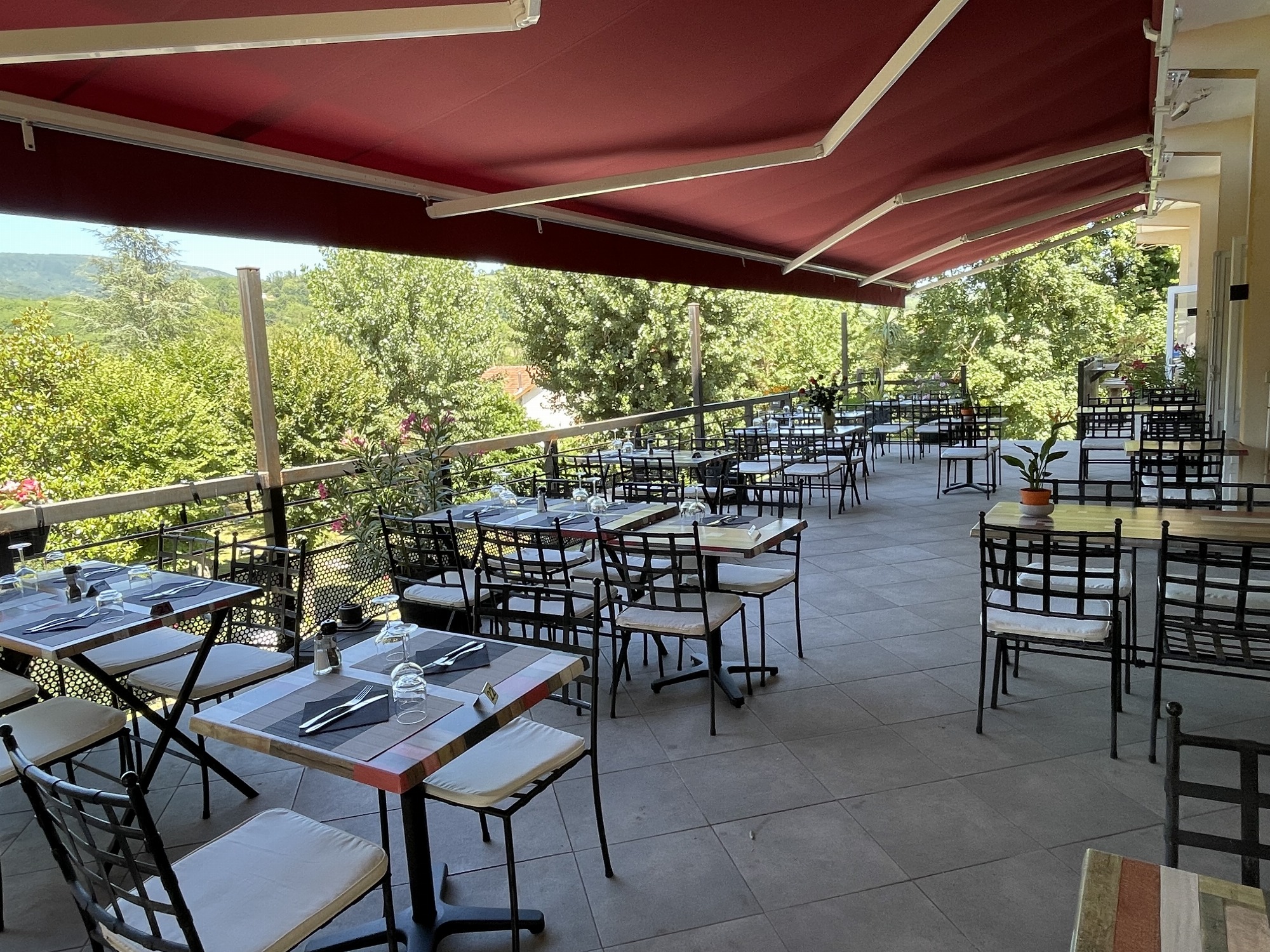 Terrasse conviviale, protégée du soleil par un large store, où une dizaine de tables sont installées, offrant une vue sur le parc arboré