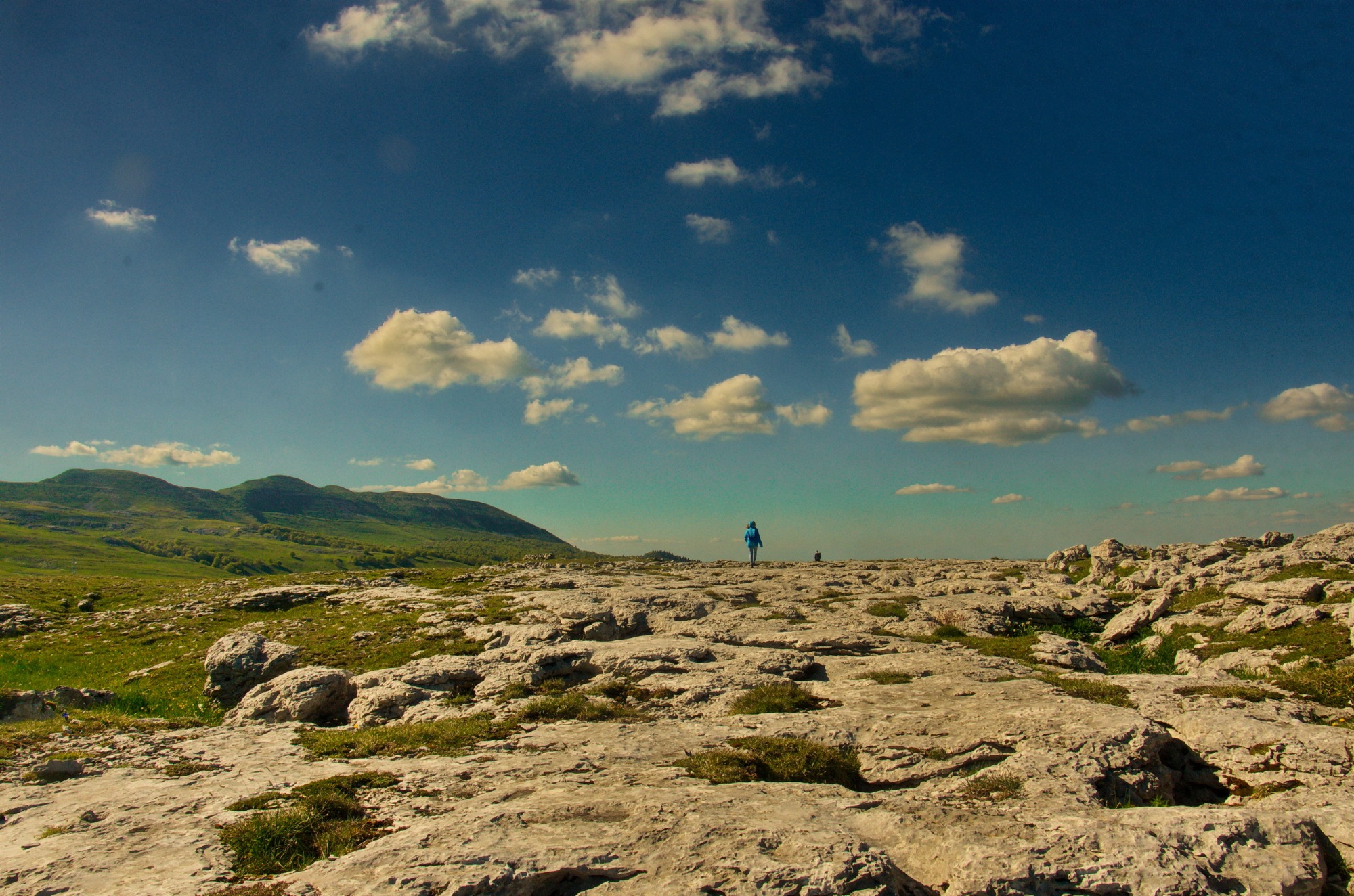 Un randonneur traverse un vaste plateau rocheux aux allures lunaires, dominé par un immense ciel bleu parsemé de nuages et entouré de collines verdoyantes.