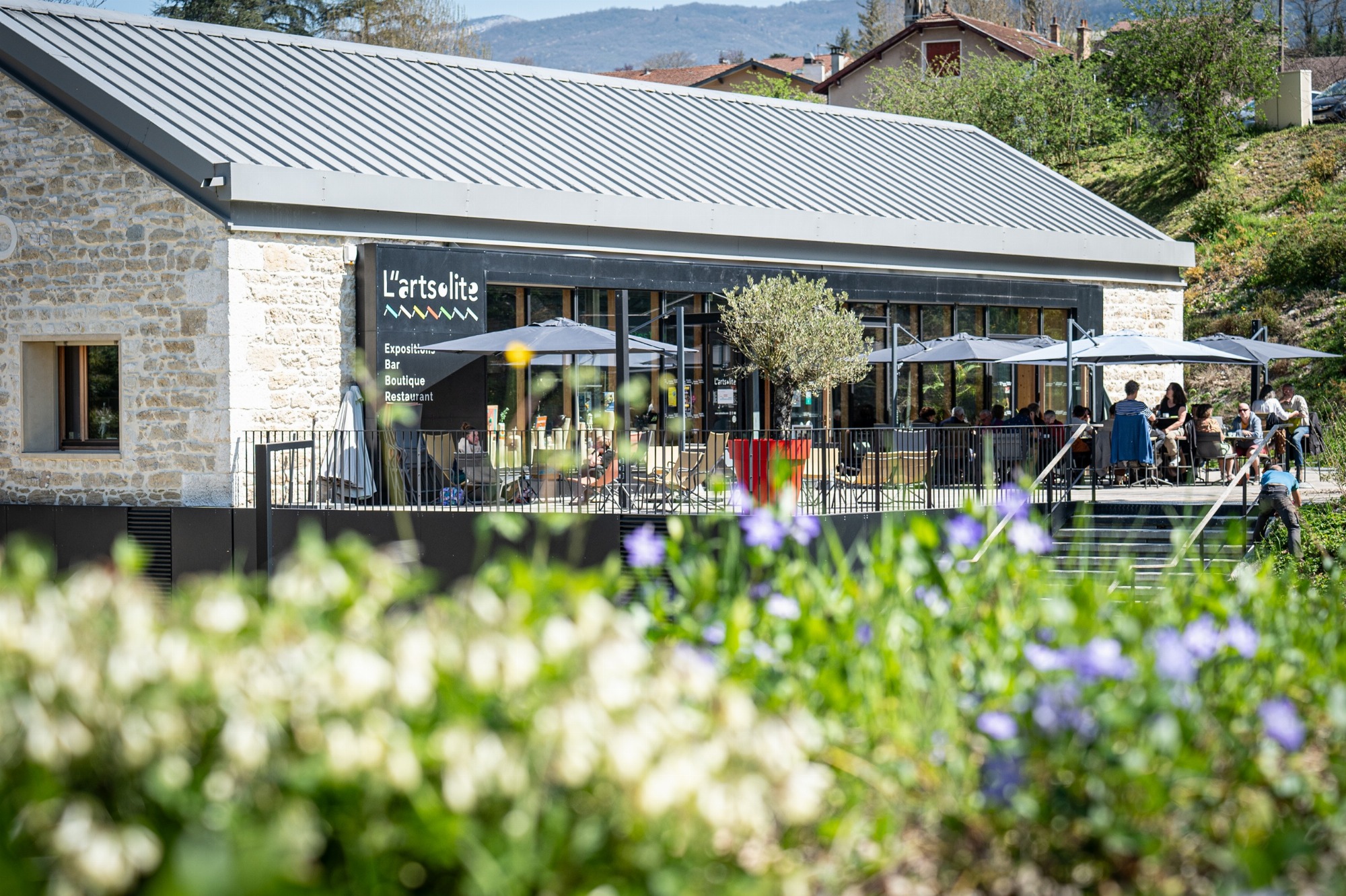 Au premier plan, des fleurs devant la terrasse où des personnes se détendent dans des transats ou partagent un moment attablées