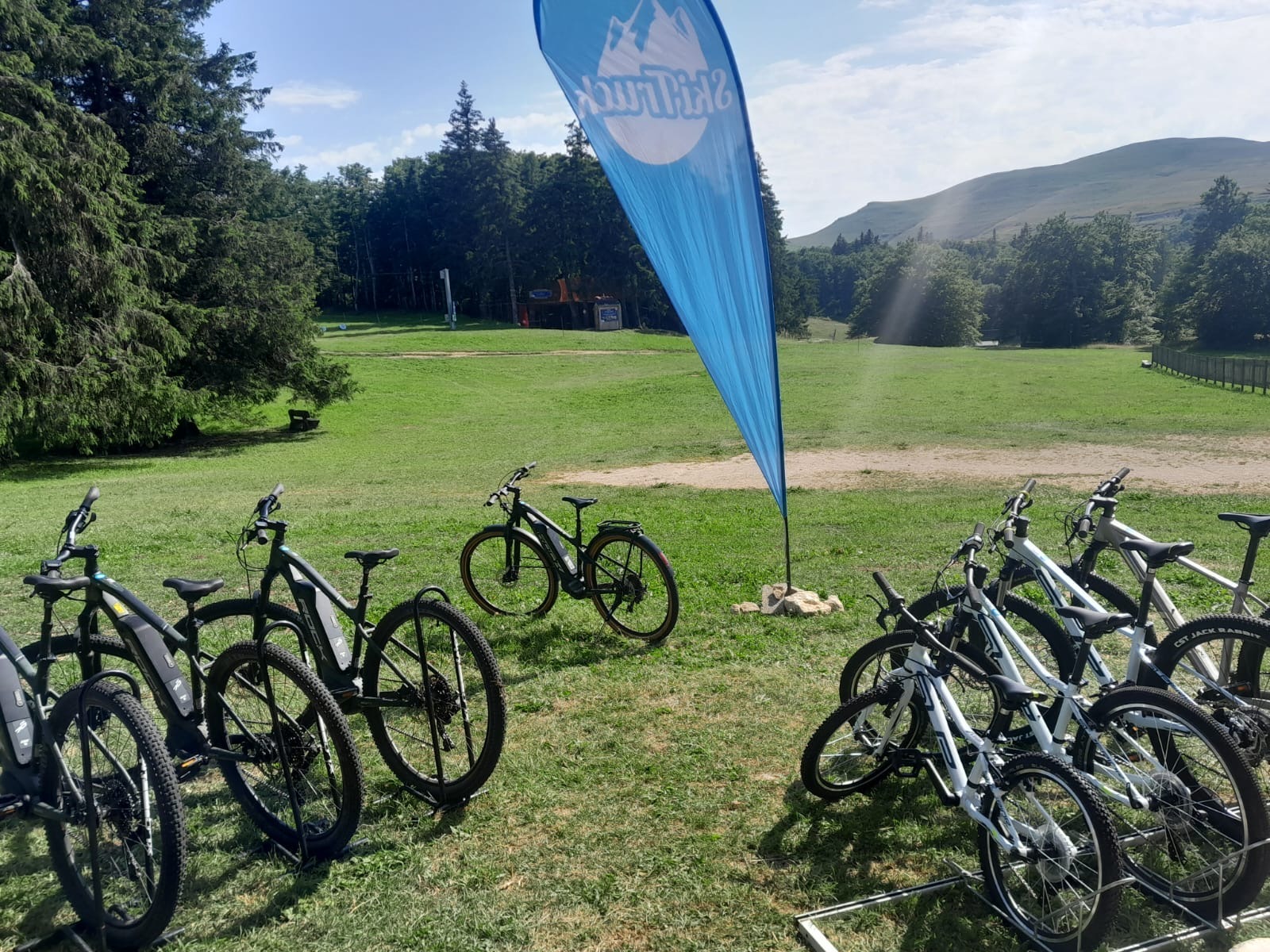 De chaque côtés de l'image des vélos et VTT à assistance électrique en présentation à l'extérieur de la boutique. Au centre, la flamme de l'entreprise.